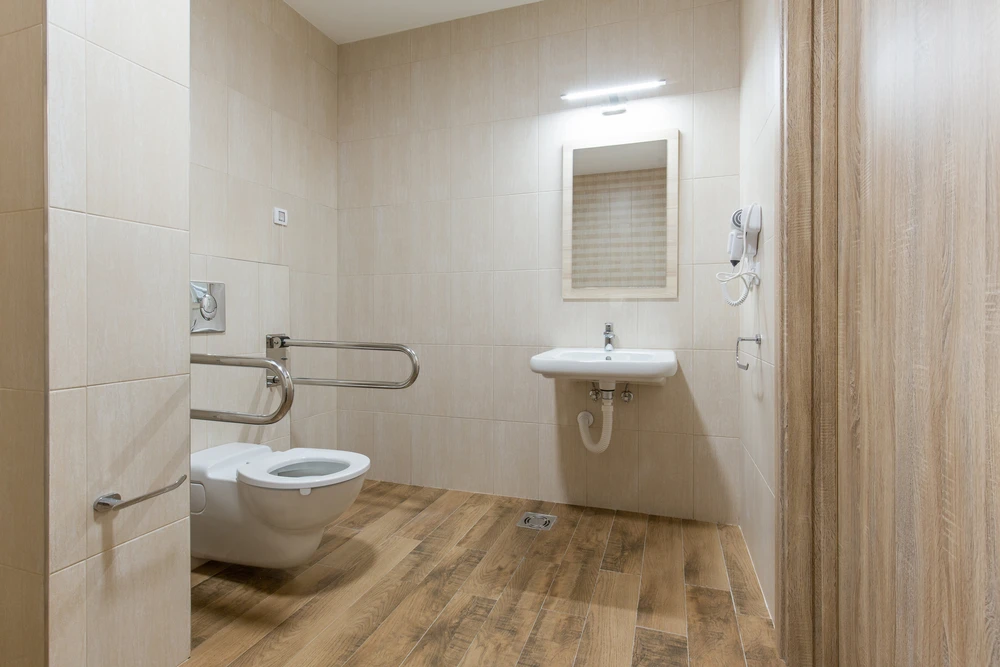Toilettes PMR dans salle de bain carrelée beige avec WC suspendu équipé de barres d'appui latérales rabattables